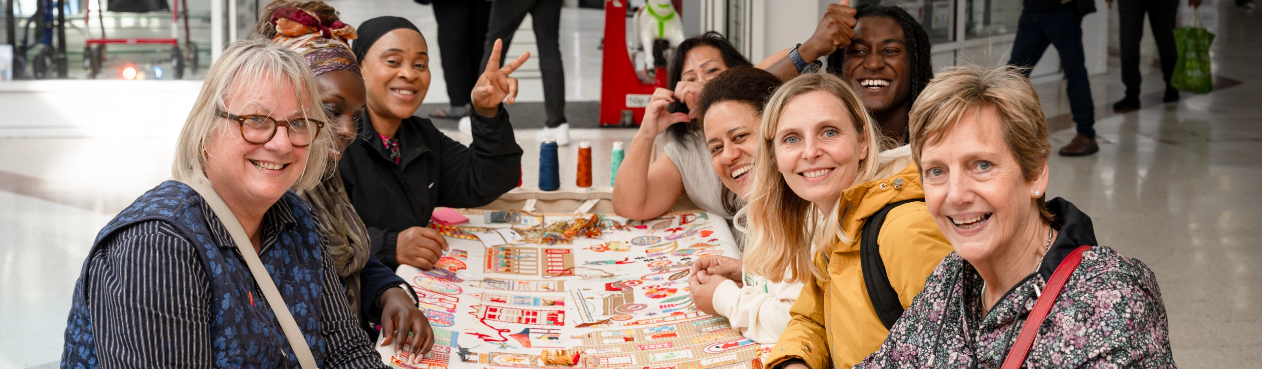 A group of 8 people, sitting around a table covered with a colourful embroidered piece of work. All 8 people are facing the camera and smiling Community involvement creating embroidery panel Creative Land Trust
