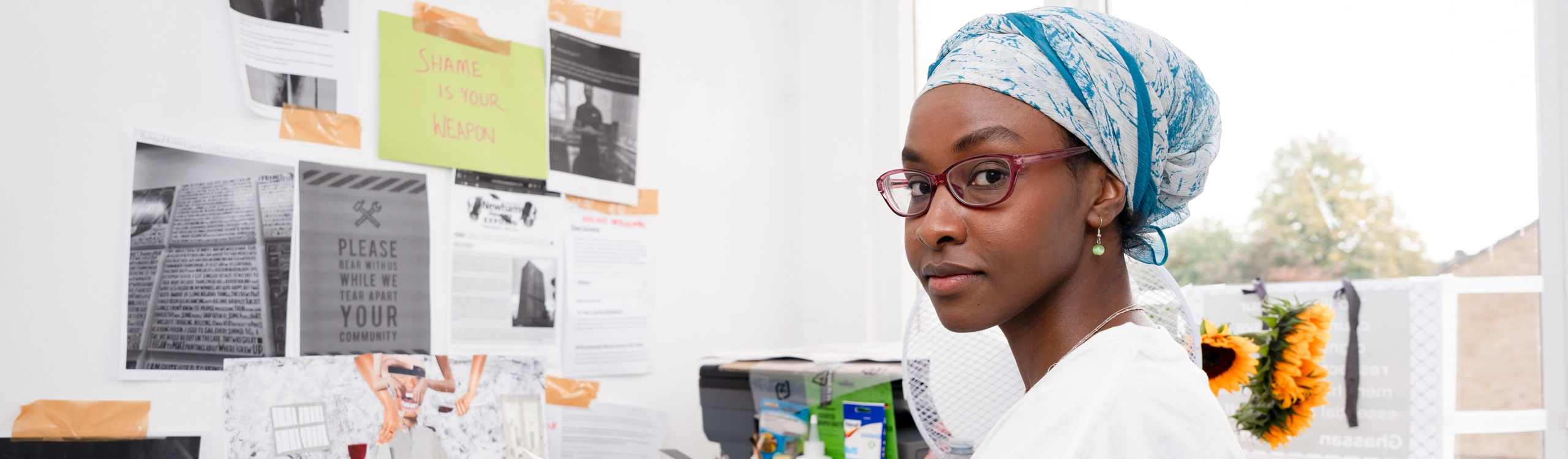 A young female artist with plum glasses and a blue and white headwrap looking over her shoulder as a pose at the camera, while at her desk. Infront of her there are posters on the wall stuck with tape Artist in her studio at Wallis Road Studio, Creative Land Trust
